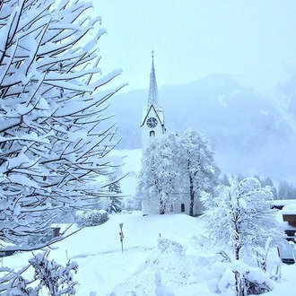 Eine verschneite Landschaft mit einer Kirche im Hintergrund. Bäume und Sträucher sind vollständig mit Schnee bedeckt.