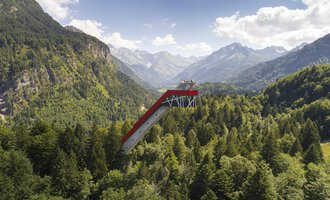 An impressive observation platform that extends above the trees, surrounded by majestic mountains and dense forests. The sky is clear and shows some clouds.