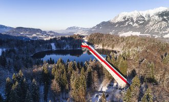 A long bridge with red and white stripes spans a tranquil, snow-covered valley and a lake. In the background, majestic mountains can be seen, glowing in the winter sun.
