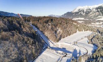 A snow-covered landscape with trees and a clear sky. In the background, mountains can be seen that complement the wintry scenery.
