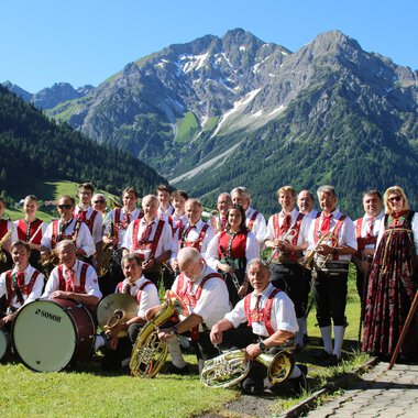Eine Gruppe von Musikern in traditioneller Tracht steht vor einer bergigen Landschaft. Sie halten Musikinstrumente und eine Fahne in der Hand. | © Musikverein d´Hirschegger | Jockl Metzger