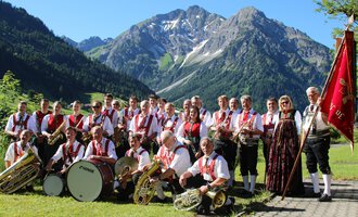 A group of musicians in traditional attire stands in front of a mountainous landscape. They are holding musical instruments and a flag in their hands. | © Musikverein d´Hirschegger | Jockl Metzger