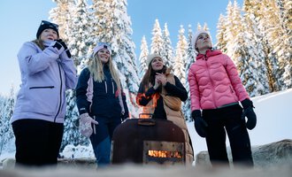 A group of women stands around a fire in a snowy landscape. In the background, tall, snow-covered trees and a clear blue sky can be seen.