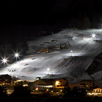 A beautifully lit ski resort at night with white snow-covered mountains. The lifts and slopes are clearly visible, while the surrounding buildings are gently illuminated.