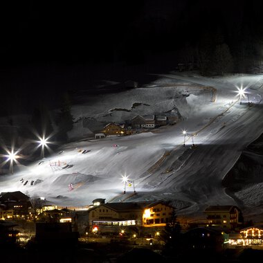 A beautifully lit ski resort at night with white snow-covered mountains. The lifts and slopes are clearly visible, while the surrounding buildings are gently illuminated.