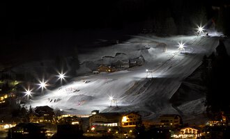 Ein wunderschön beleuchteter Skiort bei Nacht mit weißen Schneebergen. Die Lifte und Pisten sind gut sichtbar, während die umliegenden Gebäude sanft erleuchtet sind.