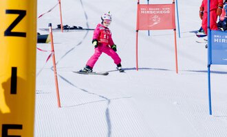 Ein kleiner Skifahrer in pinker Skiausrüstung fährt durch ein Tor auf einer verschneiten Piste. Im Hintergrund sind weitere Skifahrer und Farbmarkierungen zu sehen.