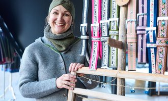 A smiling woman stands in front of a stand with colorful ribbons. She is wearing a gray sweater and a scarf.