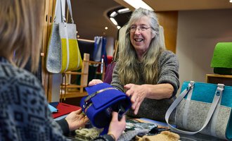 A saleswoman smiles and shows a blue bag to a customer. In the background, more bags and handicrafts are visible.