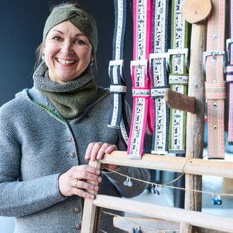 Eine freundlich lächelnde Frau steht vor einem Ständer mit bunten Bändern. Sie trägt einen grauen Pullover und einen Schal.
