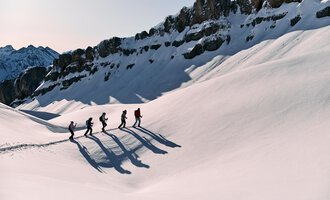 A group of hikers is walking through a snowy landscape. Majestic mountains can be seen in the background. | © Kleinwalsertal Tourismus eGen | Oliver Farys