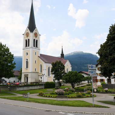 Eine malerische Kirche mit einem hohen Turm steht im Mittelpunkt eines kleinen Platzes. Im Hintergrund sind grüne Bäume und Berge zu sehen.