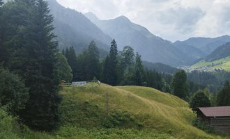 A picturesque landscape with green hills and tall mountains. In the foreground, there is a small cabin surrounded by trees. | © Nicole Lughammer