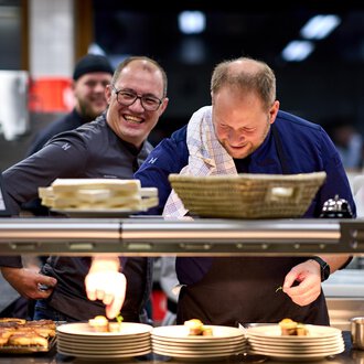 Two chefs are smiling in a kitchen as they prepare dishes. In the background, another chef can be seen, who also appears to be cheerful.