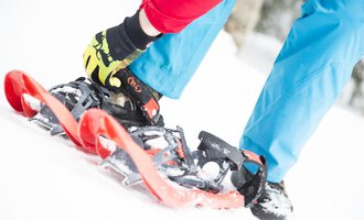A person is fastening snowshoes while standing on the white snow. The background is blurred, indicating a winter forest. | © Kleinwalsertal Tourismus | Oliver Farys