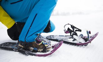 A person straps on snowshoes to hike in the snow. The surroundings are wintry and covered in snow. | © Kleinwalsertal Tourismus | Oliver Farys