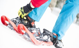 A person straps on snowshoes while standing in the snow. The ground is white and the clothing is colorful. | © Kleinwalsertal Tourismus eGen | Oliver Farys