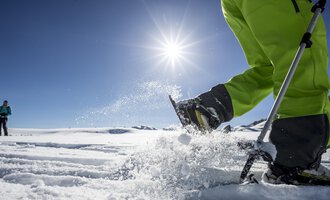 A sunny day in the snow with a person working in the snow with a ski pole. In the background, another person can be seen. | © Kleinwalsertal Tourismus | Dominik Berchtold