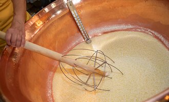 In einem großen Kupferkessel wird eine Mischung zubereitet. Ein Holzrührer wird in die goldene Masse eingetaucht.
