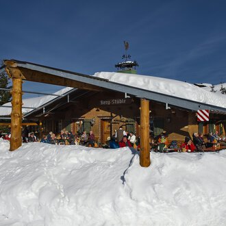 Eine gemütliche Berghütte im Schnee mit Gästen auf der Terrasse. Der Himmel ist klar und blau, und die Umgebung ist winterlich. | © Bergstüble | Michaela Beck Dornach