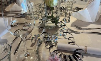 Zwei Sektgläser stoßen auf einem festlich dekorierten Tisch an. Im Hintergrund sind rote Weihnachtskugeln und goldene Dekoration zu sehen. | © Genuss- & Aktivhotel Sonnenburg