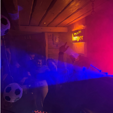 A lively party in a cozy room with colorful lights and fog.  
People are dancing and seem to be having a lot of fun.