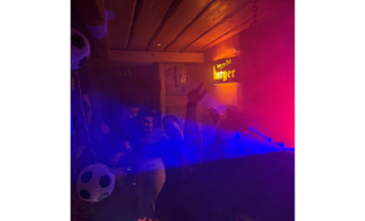 A lively party in a cozy room with colorful lights and fog.  
People are dancing and seem to be having a lot of fun.