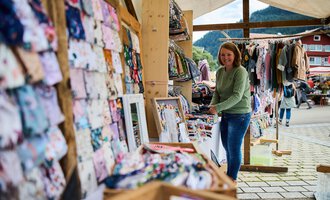 Ein Marktstand mit verschiedenen bunten Stoffen und Kleidung. Eine Frau durchsucht die Auslagen und genießt die Atmosphäre. | © Kleinwalsertal Tourismus | Fotograf Oliver Farys