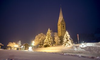 Eine verschneite Landschaft bei Nacht mit einer Kirche und beleuchteten Bäumen. Der Himmel ist dunkelblau und die Straße ist sichtbar. | © Kleinwalsertal Tourismus | Dominik Berchtold