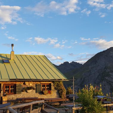 Eine gemütliche Berghütte mit grünem Dach in den Alpen. Im Hintergrund erstrecken sich majestätische Berge unter einem blauen Himmel. | © Alpe Kuhgehren | Lisa Hiesinger