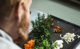 A chef stands at a table with fresh herbs and vegetables. The ingredients are colorful and ready for preparation. | © Alpenhotel DAS KÜREN