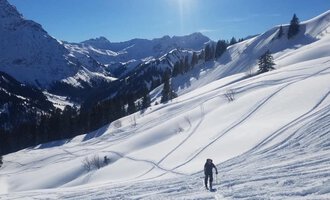 A skier stands on a snow-covered landscape with tall mountains in the background. The sky is clear and blue sunshine shines on the scene.