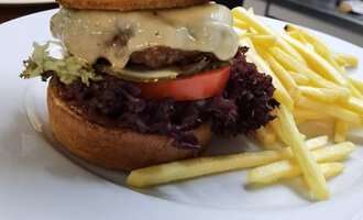 Ein großer Burger mit zwei Brötchen, einem Fleischpatty, Käse, Salat und Tomate. Dazu sind Pommes Frites auf dem Teller.