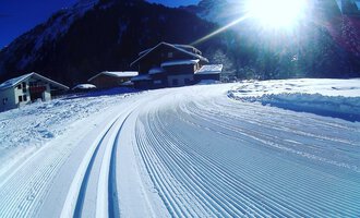 Eine verschneite Landschaft mit präparierten Skispuren und einem sonnigen Himmel. Im Hintergrund sind Hütten und Berge zu sehen.