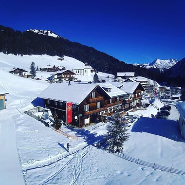 A snow-covered landscape with a cozy wooden house and mountains in the background. The sky is clear and blue.