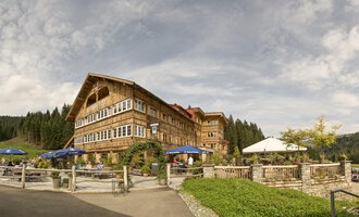 A rustic hotel with a wooden facade in a beautiful natural landscape. The outdoor area has seating and sun umbrellas under a cloudy sky. | © Auenhütte | Yorck Dertinger