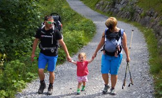 Eine Familie wandert auf einem schmalen Weg in der Natur. Die Eltern halten die Hände ihrer kleinen Tochter und tragen Rucksäcke. | © Austrian Ski- & Service Ranch | Trixi Leitner