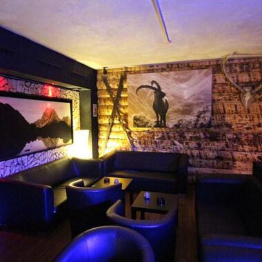 A cozy interior with dark leather furniture and wooden paneling. On the wall hangs a picture of a mountain panorama and a stylized goat motif. | © Heiko Möller m&m bar
