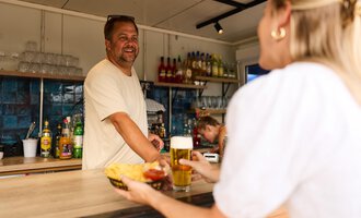 A cheerful bartender smiles at his customers at the counter. In the foreground, a portion of fries is served with a drink. | © Kleinwalsertal Tourismus | Oliver Farys