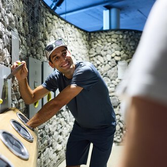 A man is smiling and interacting with a game or installation wall. The surroundings consist of stones and modern design. | © Kleinwalsertal Tourismus |  Oliver Farys