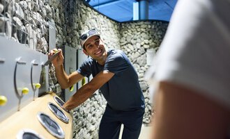 A man is smiling and interacting with a game or installation wall. The surroundings consist of stones and modern design. | © Kleinwalsertal Tourismus |  Oliver Farys