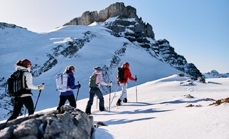 Eine Gruppe von Wanderern bewegt sich durch eine schneebedeckte Landschaft. Im Hintergrund sind beeindruckende Felsen und ein klarer blauer Himmel zu sehen. | © Kleinwalsertal Tourismus | Oliver Farys