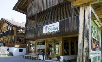 Ein Holzgebäude mit einem Schild, das „BioHof“ anzeigt. Daneben stehen Tische und Stühle im Außenbereich, umgeben von weiteren traditionellen Gebäuden. | © Kleinwalsertal Tourismus