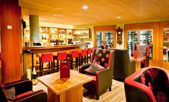 Eine gemütliche Bar mit einem Holzinterieur und roten Stühlen. Die Beleuchtung ist warm und einladend, mit Regalen voller Getränke im Hintergrund. | © Hotel Birkenhöhe | Fotograf Krug