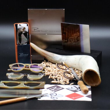 A stylish arrangement of glasses, wooden pieces, and tools. In the background, elegant packaging and a large horn bowl can be seen. | © Bräuer Optik & Akustik | AlpenMedia-Design