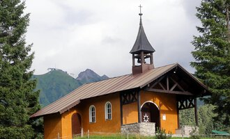 A small, orange church stands surrounded by green trees. In the background, mountains and a cloudy sky can be seen. | © Bruder Klaus Kapelle | Monika Gmeiner