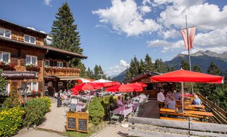 Ein gemütliches Restaurant mit Terrasse, umgeben von Natur und Bergen. Die Gäste sitzen unter roten Sonnenschirmen und genießen die Aussicht. | © Bühlalpe | Frank Drechsel