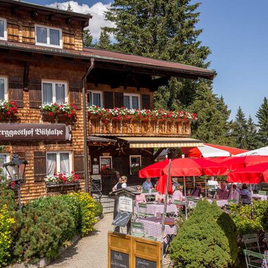 A charming inn with a wooden facade and blooming flowers. Outside, tables are set up with red umbrellas. | © Bühlalpe | Frank Drechsel