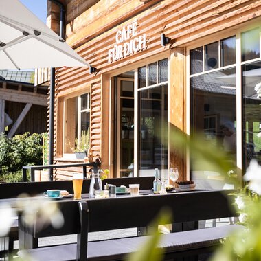 An inviting café with a wooden facade and a sunny outdoor area. Tables are set up for guests to enjoy the fresh air.
