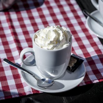 Eine Tasse mit Sahne auf einem weißen Unterteller. Der Hintergrund ist ein rot-weiß kariertes Tischläufer. | © Kleinwalsertal Tourismus | Dominik Berchtold